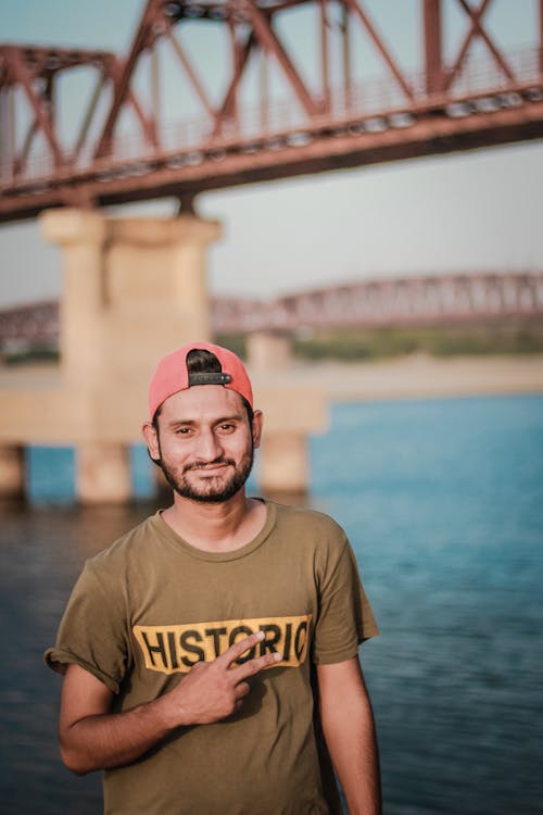 Hombre Vestido Con Camiseta Gris Con Estampado Histórico Y Gorra Roja Snapback Tomando Fotos Junto Al Cuerpo De Agua