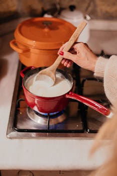 A person stirs milk in a red saucepan on a kitchen stove. Warm, cozy atmosphere.