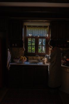 A person in a dimly lit kitchen standing by the sink with natural light coming through the window.