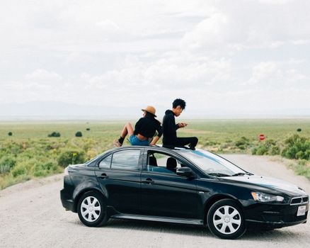 Two young adults sitting on a car roof in a scenic open landscape, symbolizing freedom.