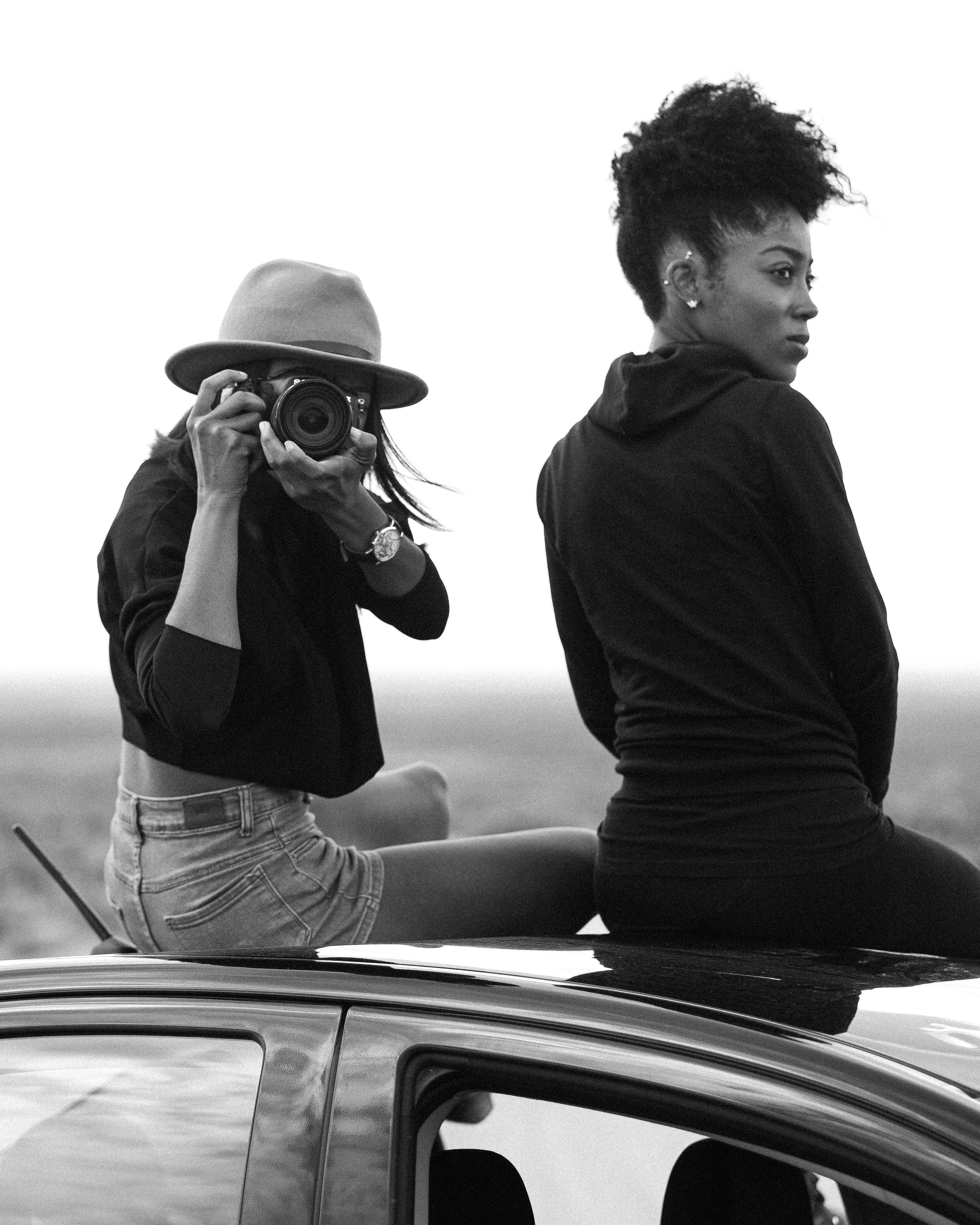 Two women enjoy a road trip, with one capturing the moment on camera in a stylish monochrome setting.