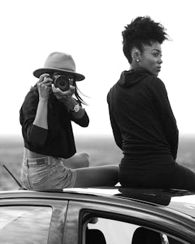 Two women enjoy a road trip, with one capturing the moment on camera in a stylish monochrome setting.