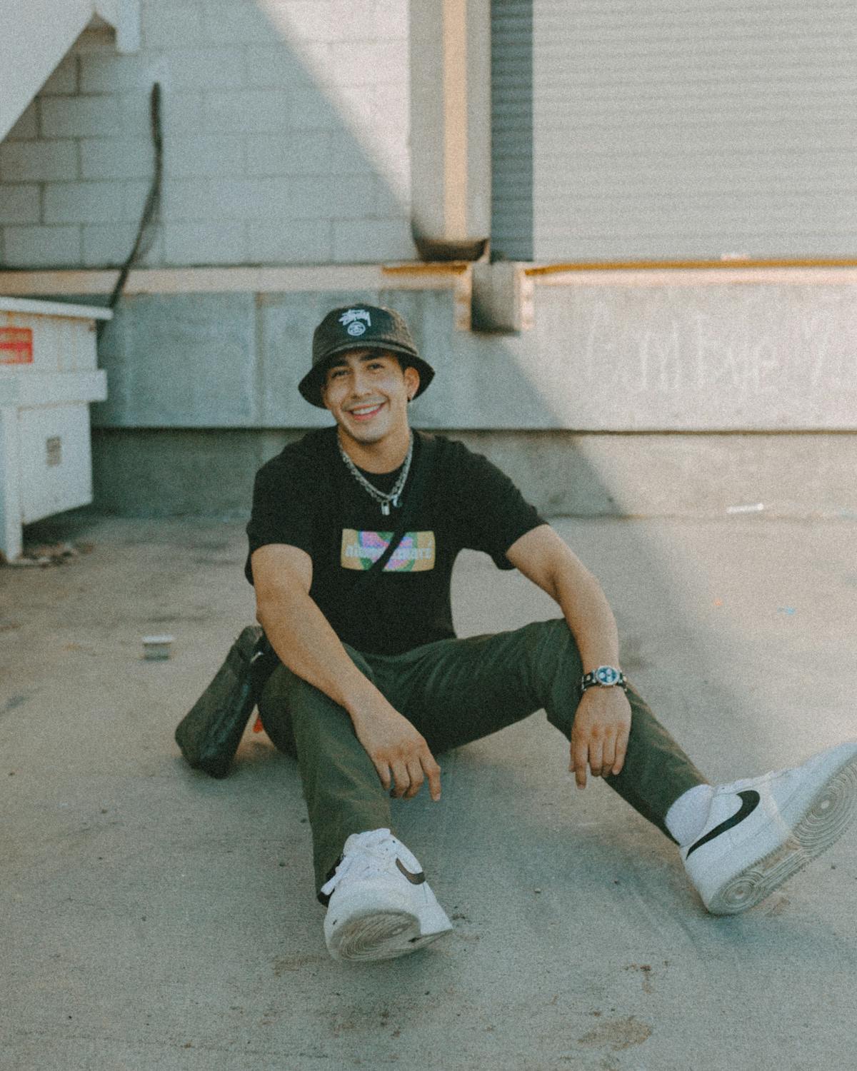 young man sitting on the ground in street fahsion clothes and a bucket hat smiling