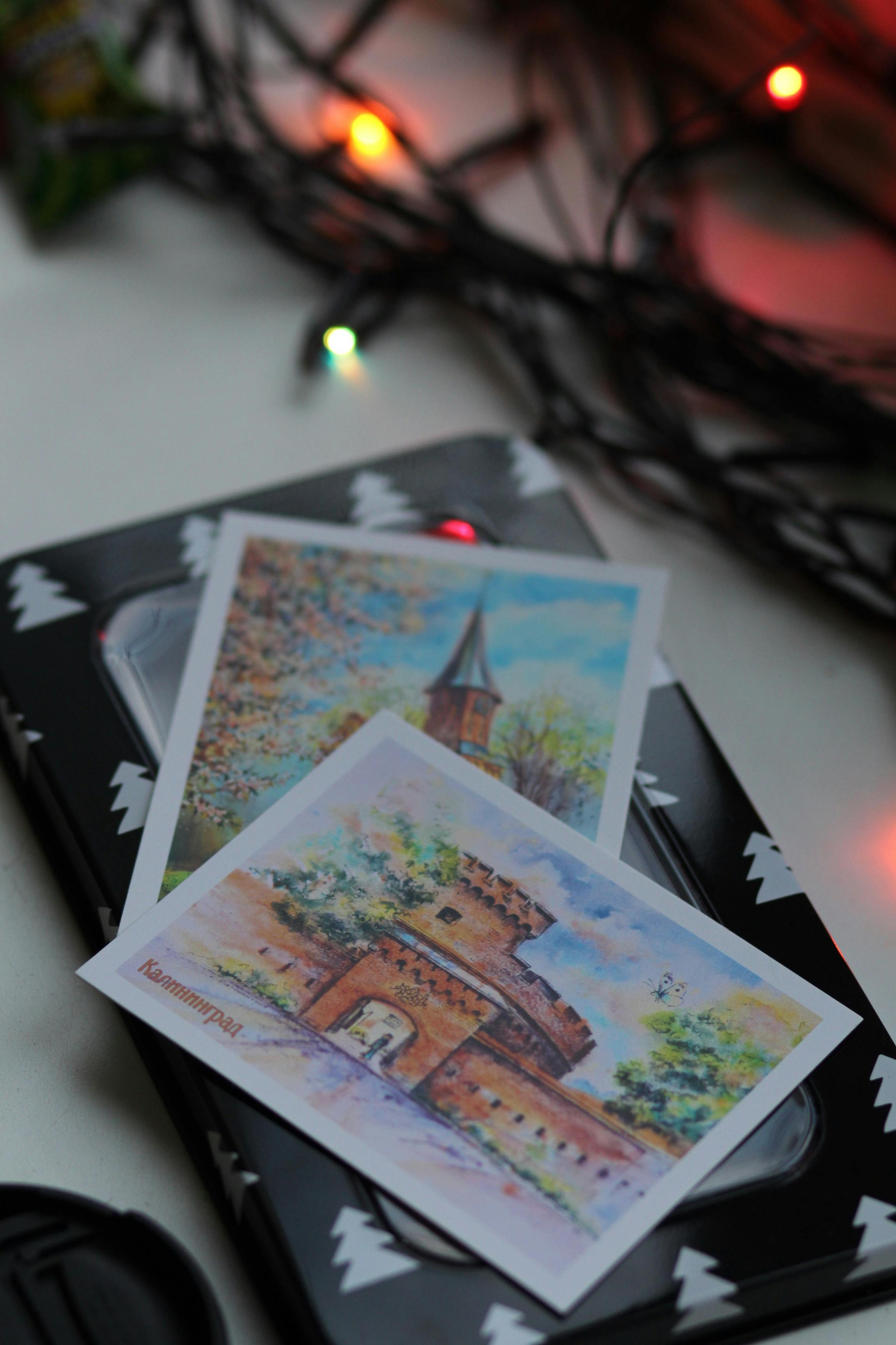 Two watercolor postcards displayed with colorful Christmas lights, creating a festive mood.