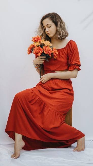 Elegant woman in a long red dress sitting with a bouquet of orange roses against a white background.