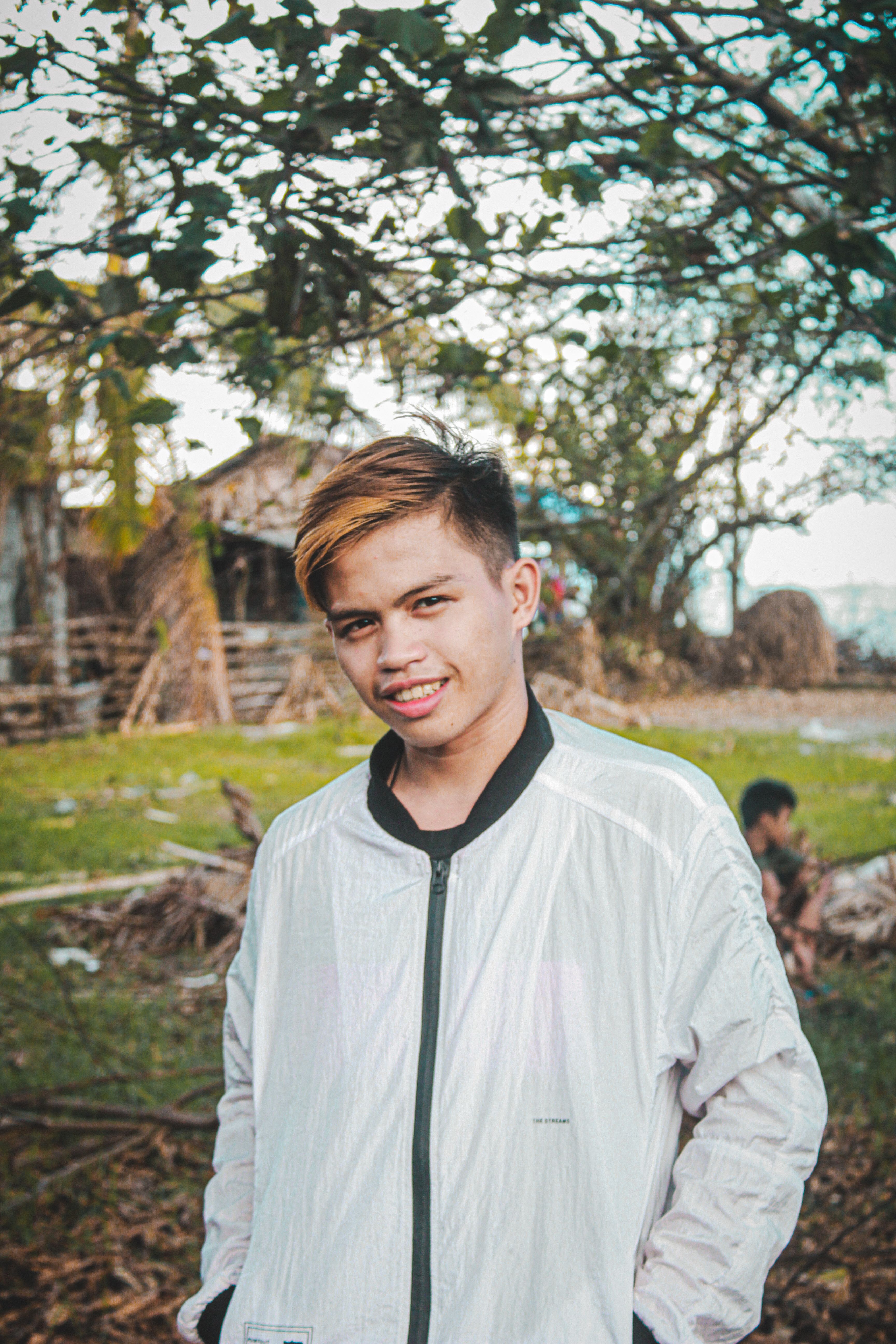 Smiling young man wearing a casual white jacket, standing outdoors with greenery in the background.