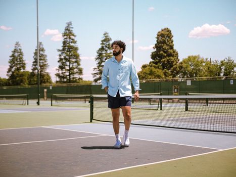 A man walks confidently on a sunny tennis court, wearing casual attire.
