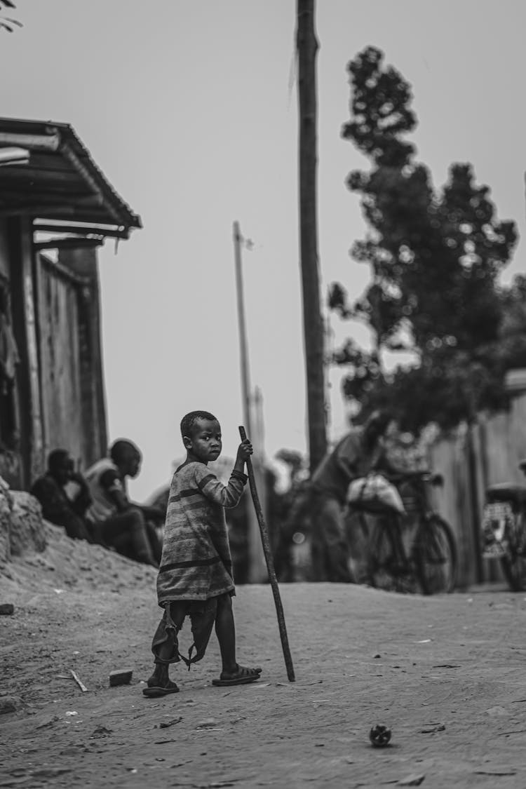 A Boy In Striped Shirt Holding A Long Stick Walking On Dirt Ground