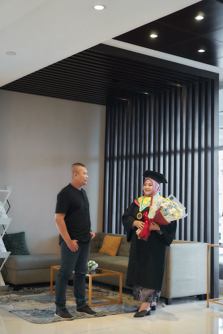 Young Woman In A Graduation Gown Holding Flowers And A Man Next To Her