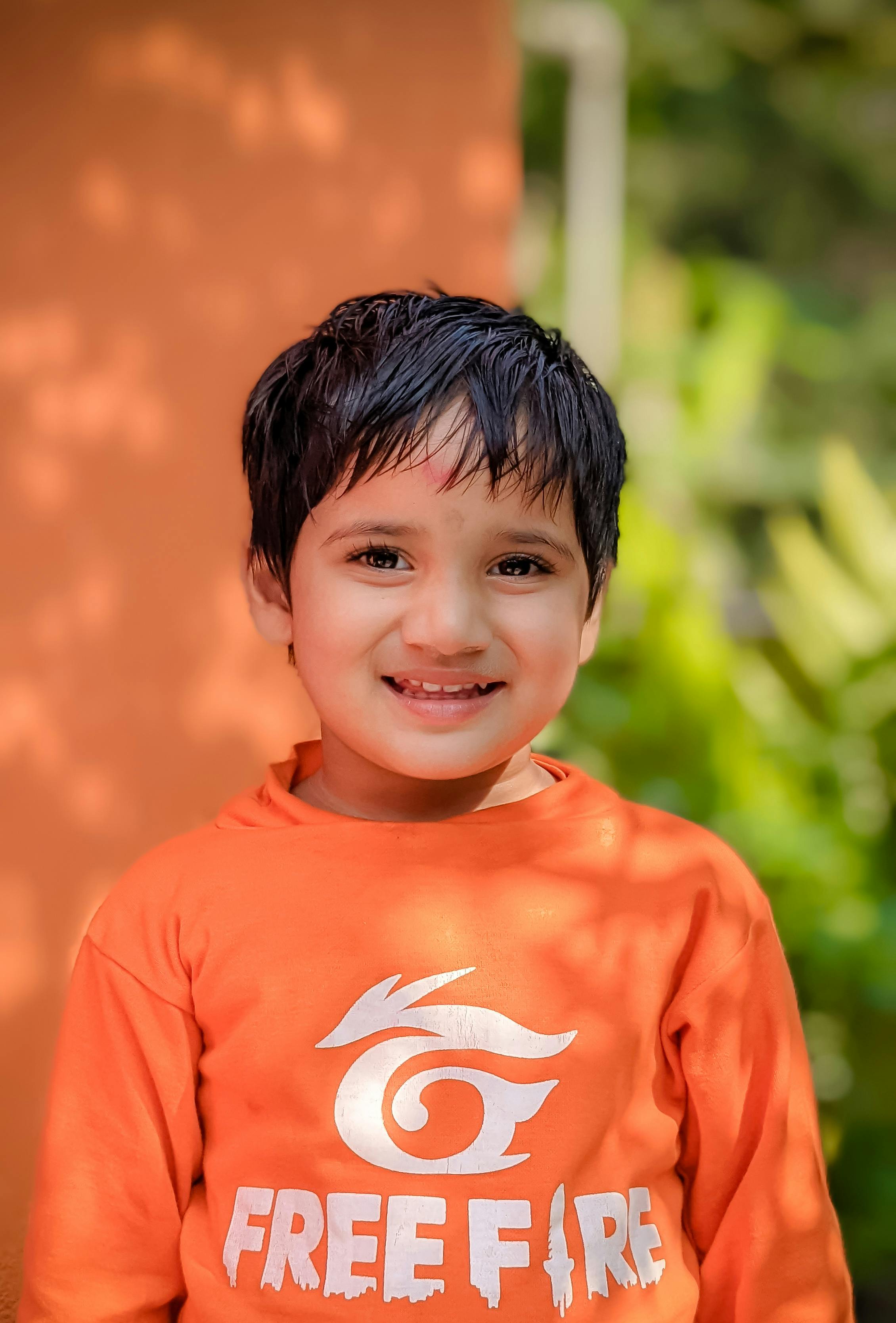 A happy child in a vibrant orange shirt smiles warmly outdoors, surrounded by green foliage.