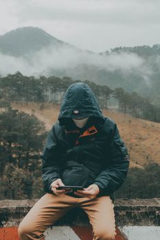 A solitary figure wearing a hoodie sits outdoors using a smartphone, surrounded by a misty forest landscape.