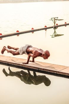An elderly man practicing advanced yoga pose on a boardwalk near the water.