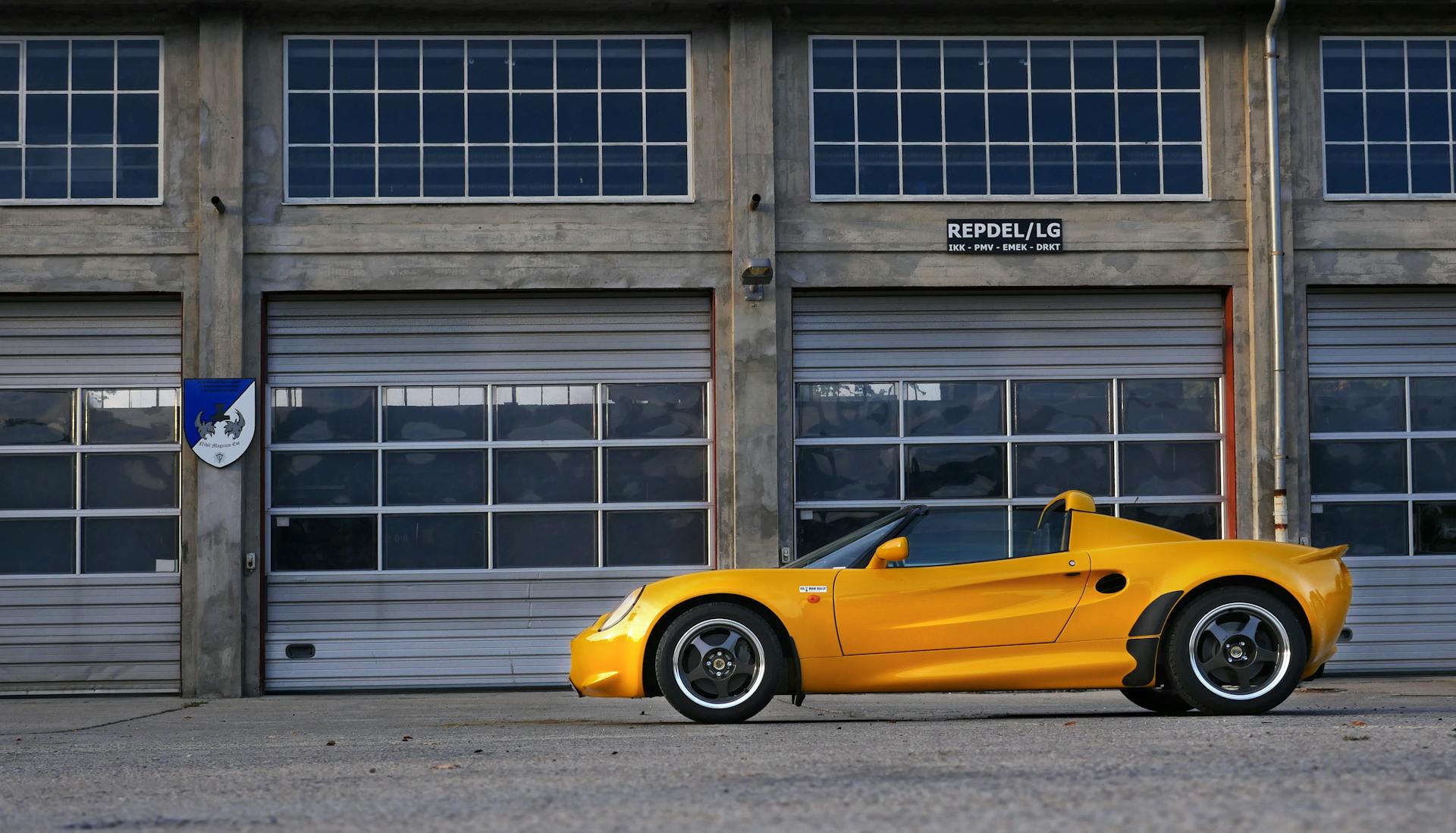 A vibrant yellow classic car parked in front of industrial garage doors on a sunny day.