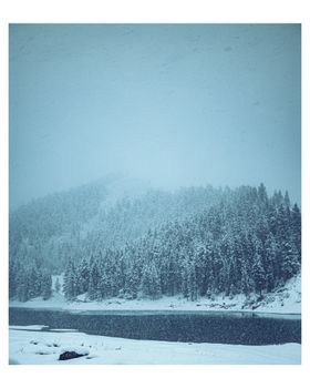 A serene winter scene of a snow-covered forest by a river with a misty, cold atmosphere.