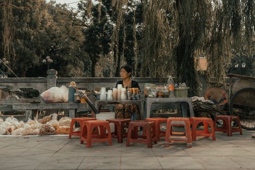 A woman sits at an outdoor street food stall surrounded by snacks and drinks. Rustic vibe.