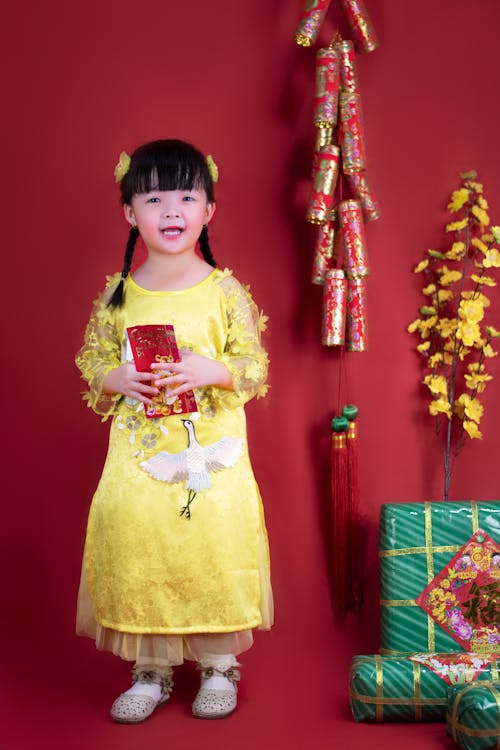 Young girl in a yellow traditional dress smiling and holding a red envelope, standing against a red background decorated with Lunar New Year ornaments