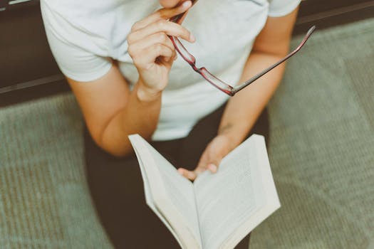 Young woman sitting indoors, reading a book while holding eyeglasses. A moment of focus and reflection.