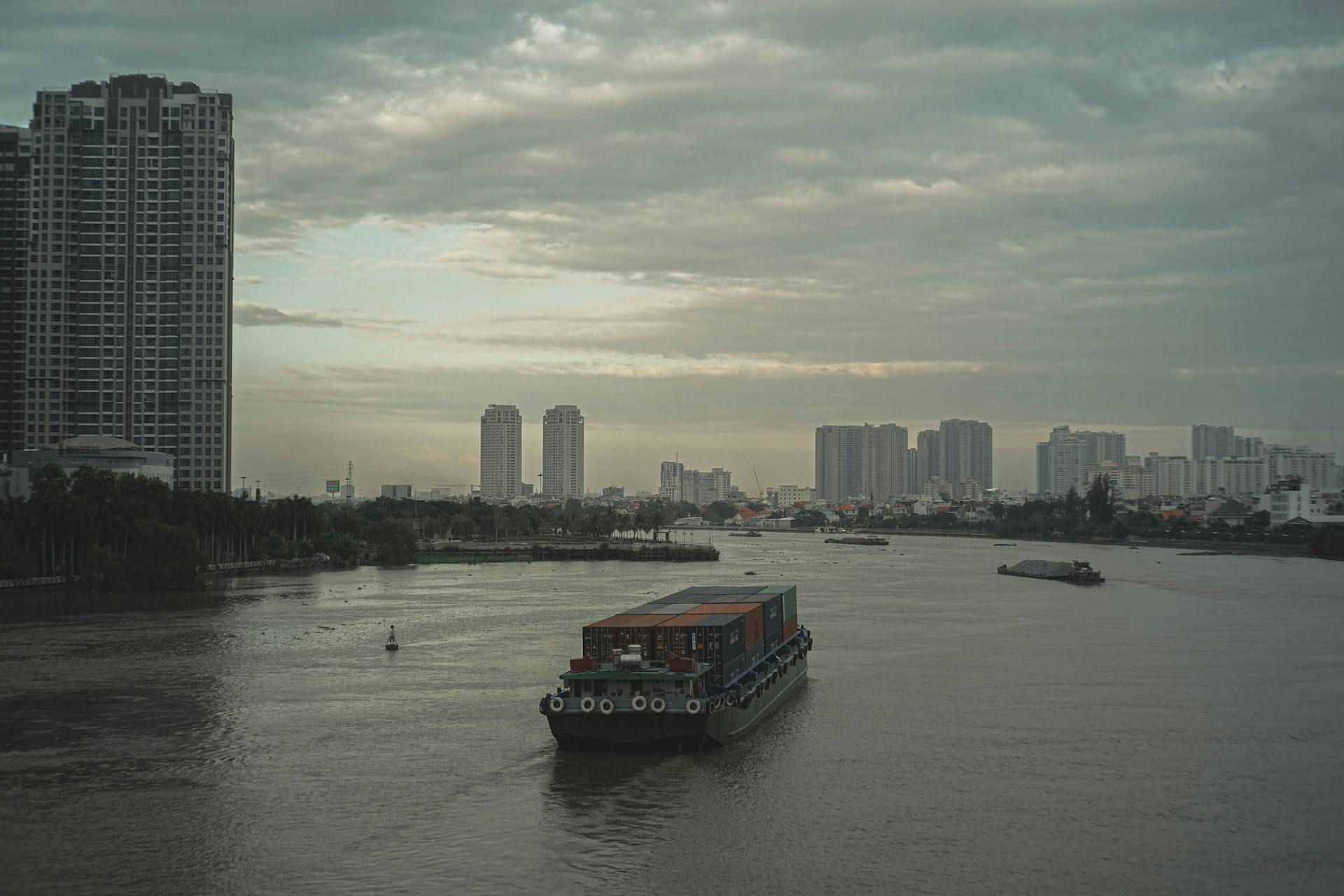 A container ship sails on a river against a backdrop of urban buildings under a cloudy sky.