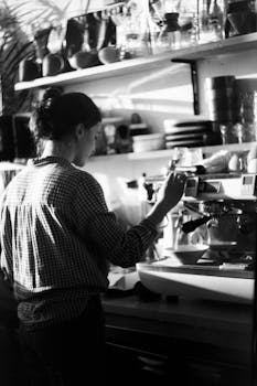 Black and white photo of a woman making coffee in a cozy cafe environment, highlighting artisanal brewing.