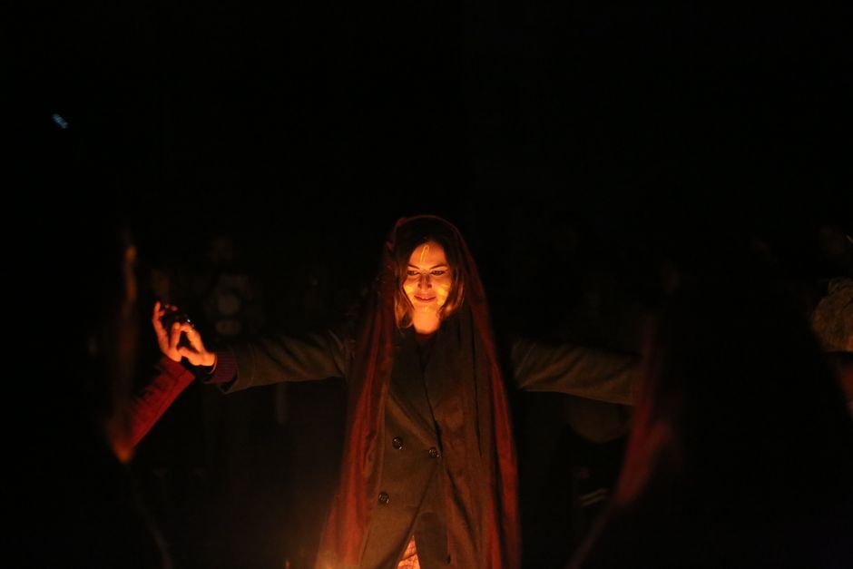 A woman performs a mystical ritual around a fire at night in Italy, surrounded by people.