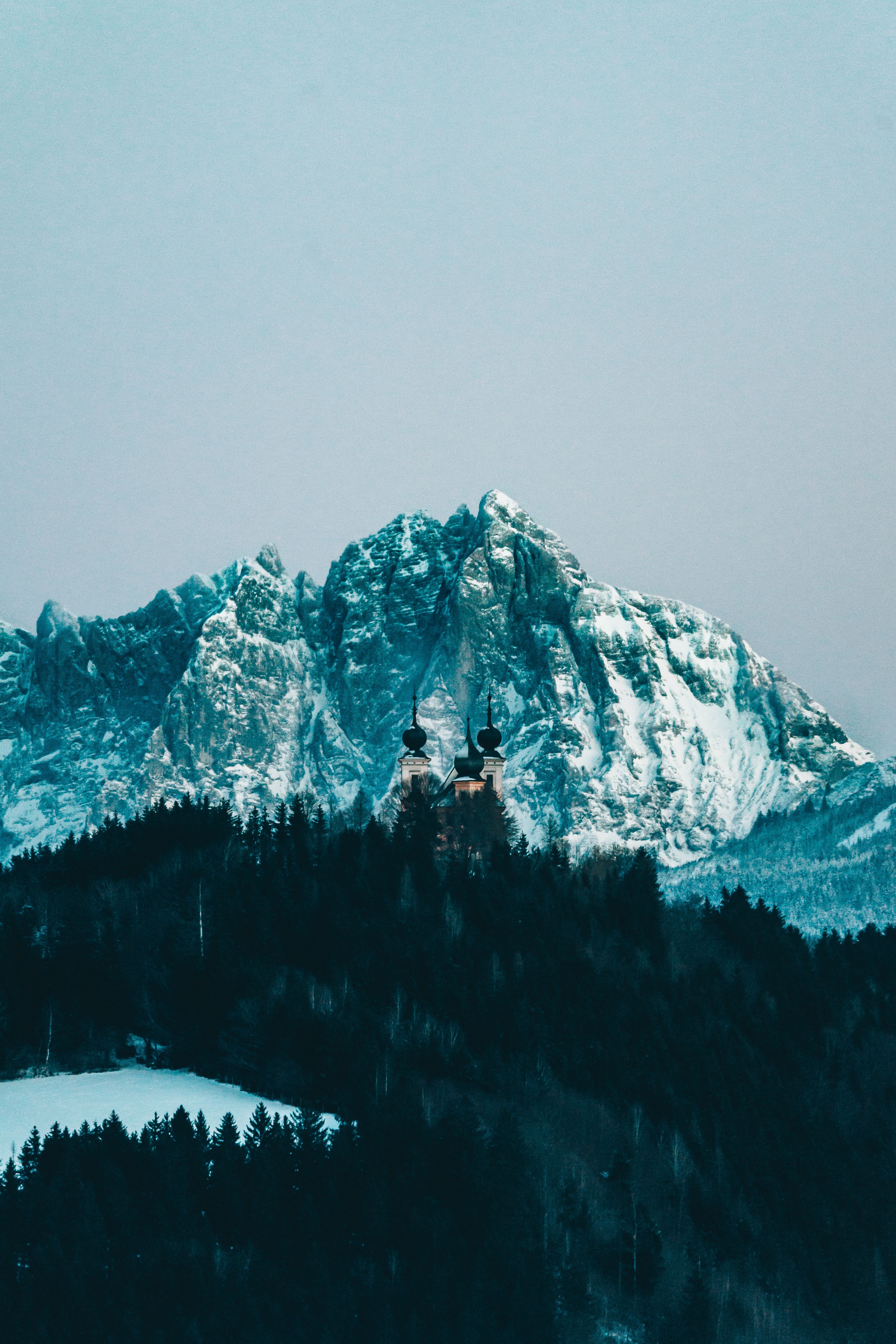 Snow-covered mountains with a church amid dense forest under a gloomy winter sky.