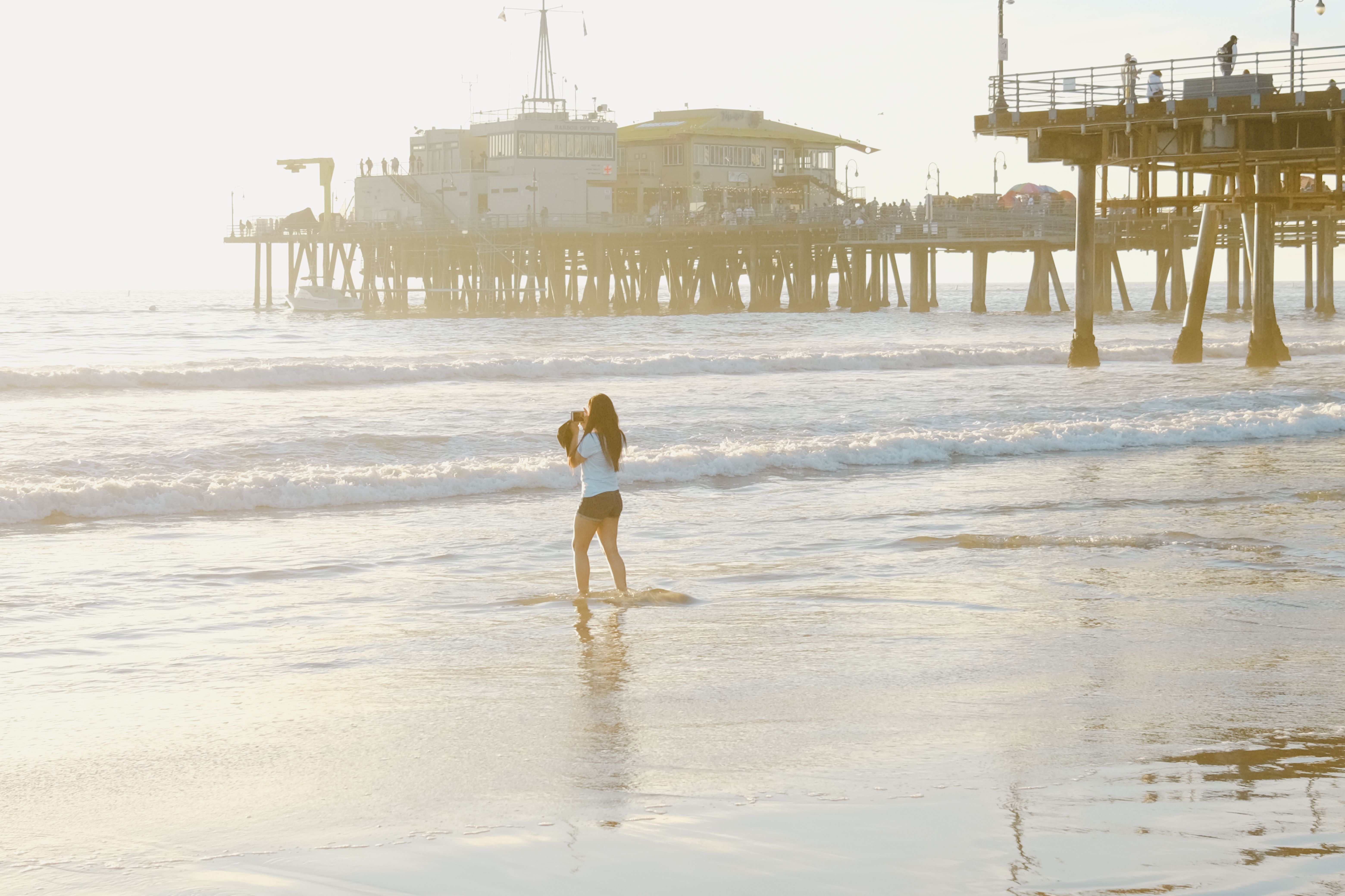 "Woman in a casual outfit standing at the shore with Santa Monica Pier in the background on a sunny beach day"