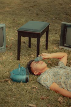 A young man lying on grass surrounded by vintage electronics, embracing nostalgia.