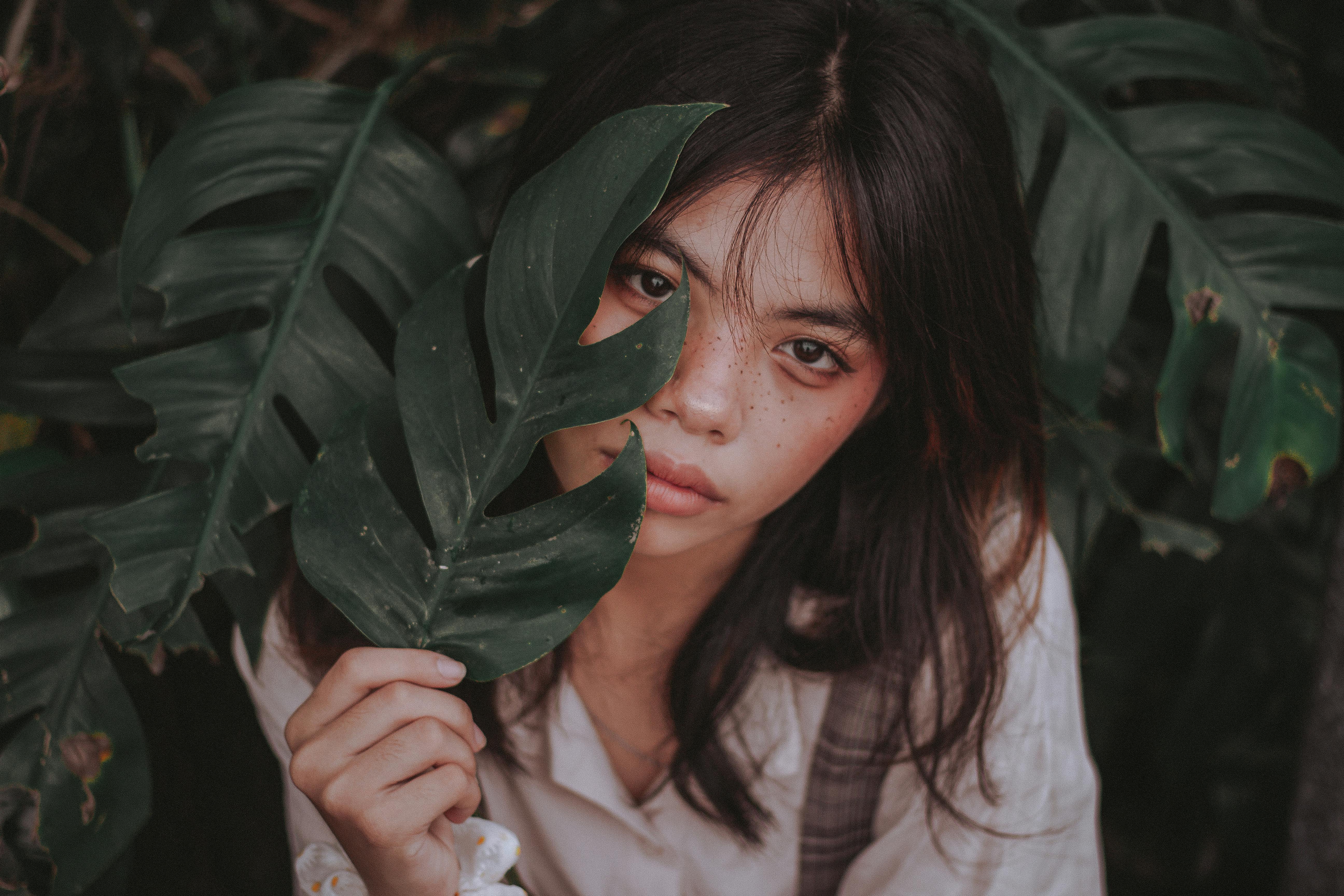 A woman holding a Monstera leaf looks directly at the camera in a lush outdoor setting.