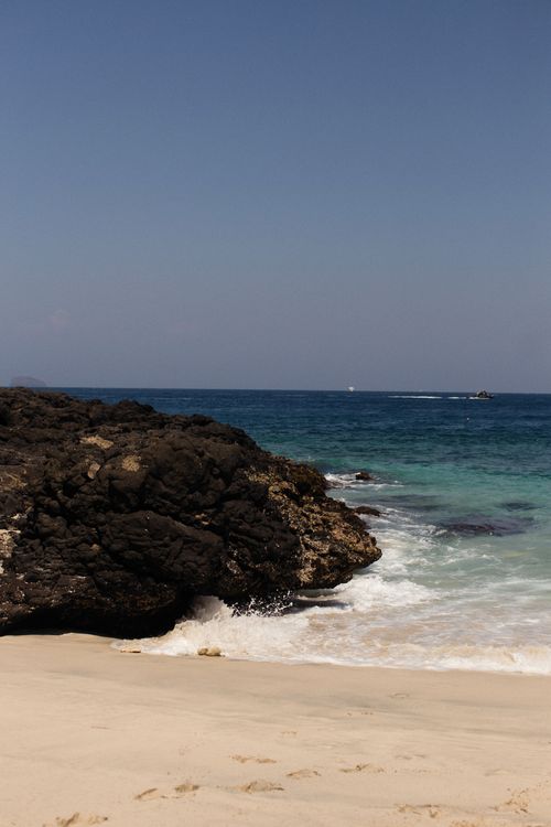 Free A tranquil beach scene with rugged rock formations and gentle waves lapping the shore under a clear blue sky. Stock Photo
