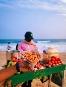Hand holding a cone of bhel puri in front of a beachside snack vendor with ocean in background.