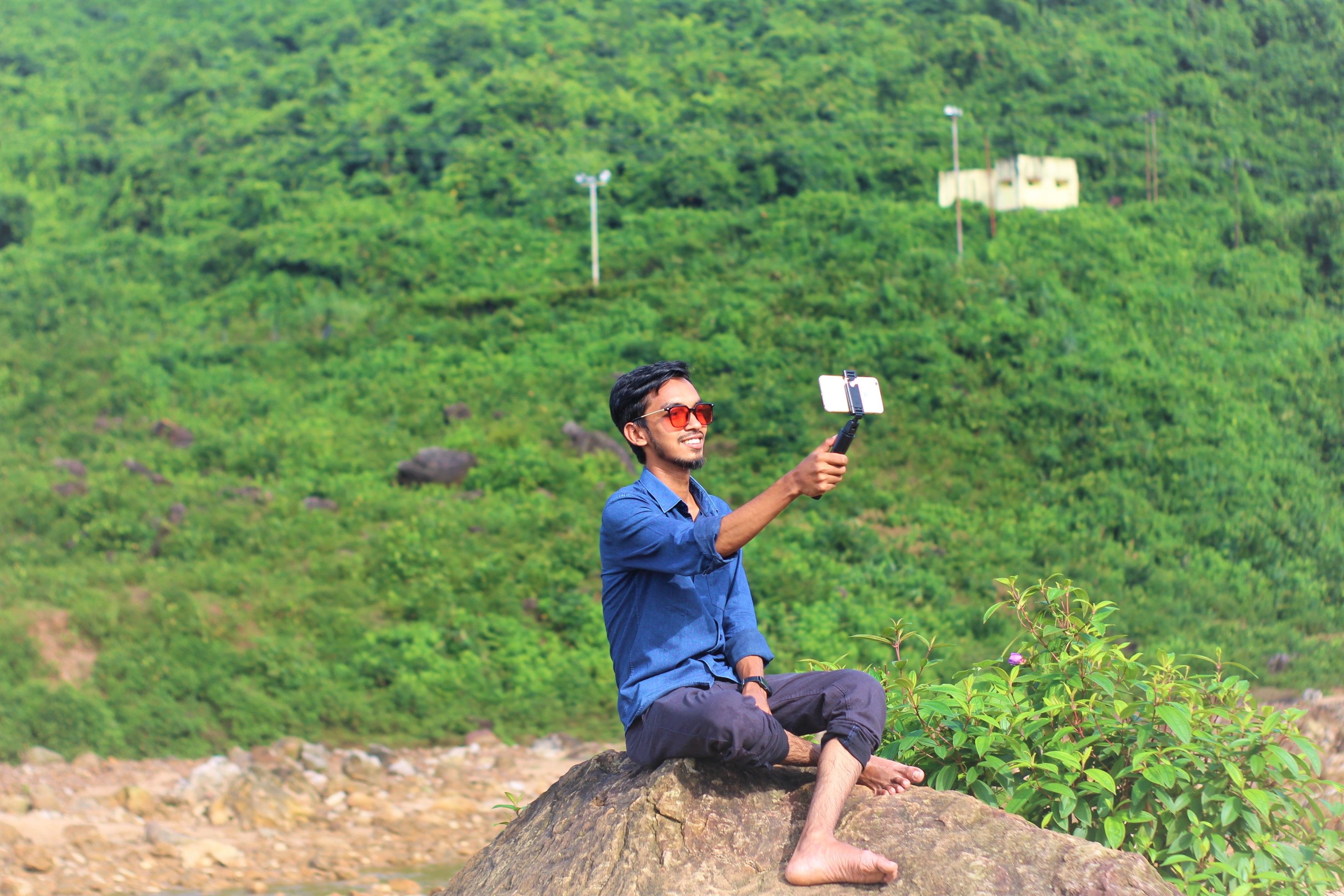 A smiling man sits on a rock taking a selfie with lush green hills in the background.