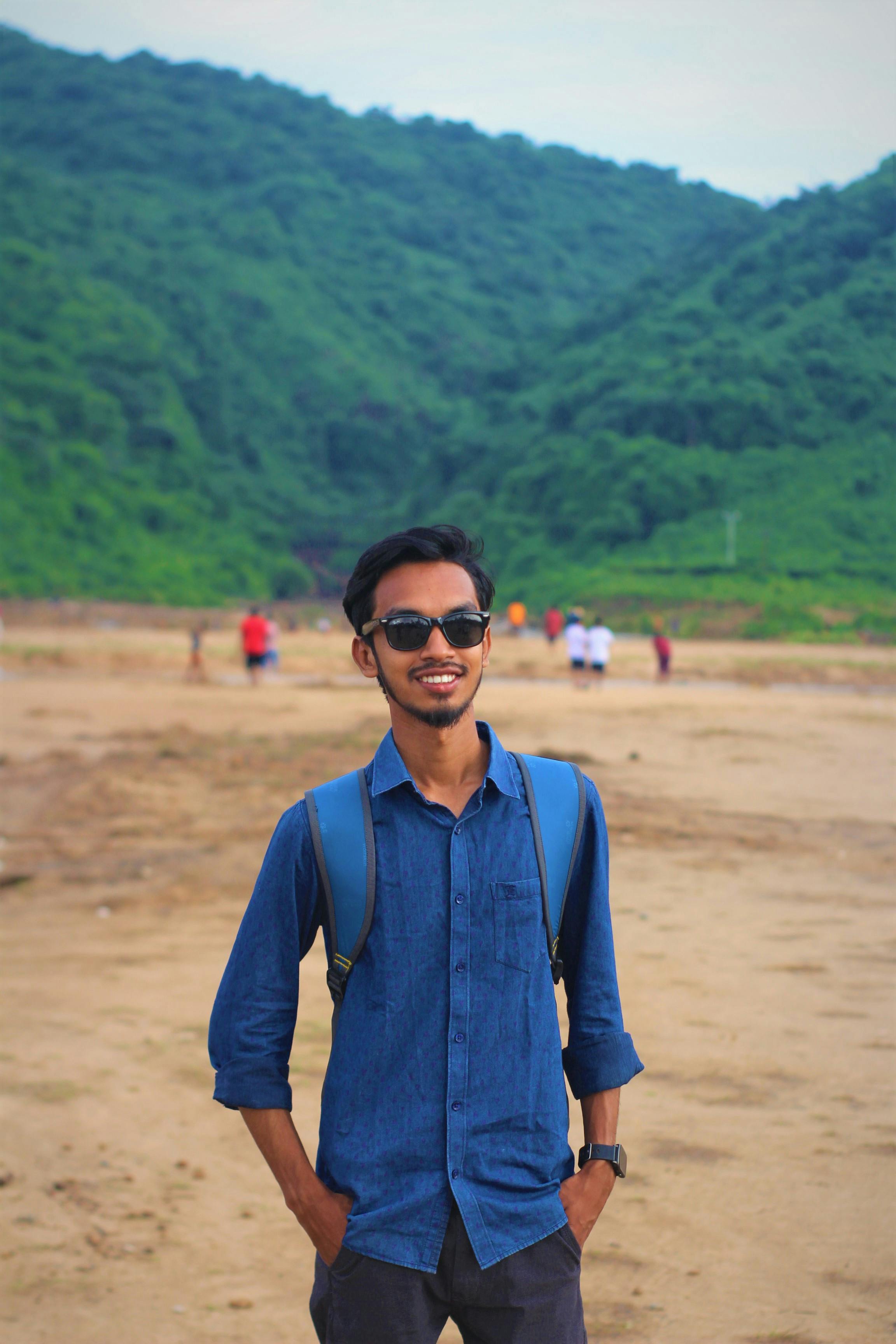 Casual portrait of a smiling man in sunglasses and backpack by the beach with lush green hills in the background.