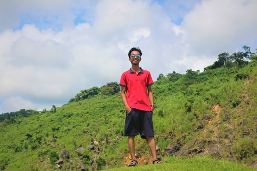 Casual man in sunglasses smiling outdoors on a sunny day with green hill in the background.