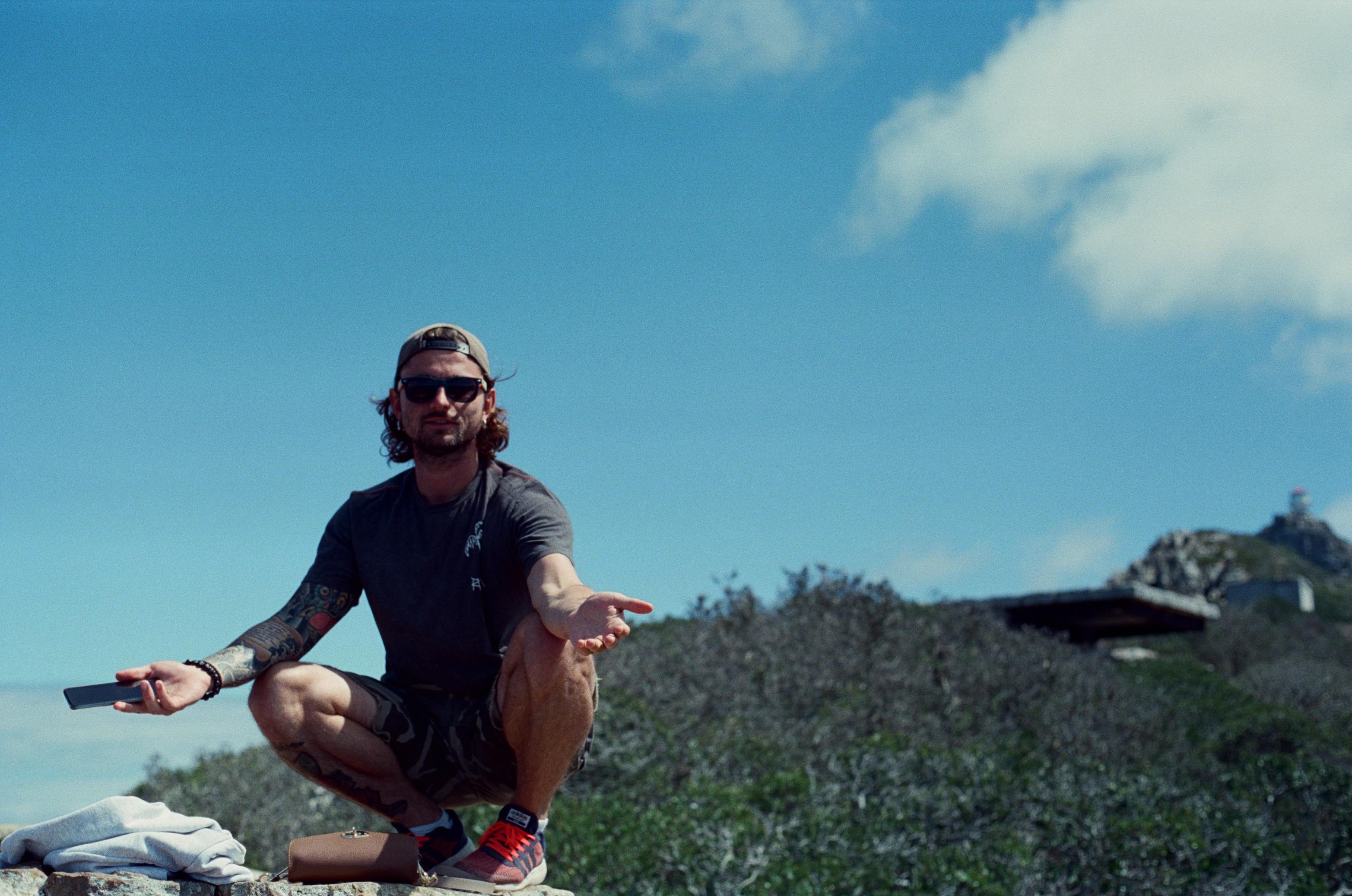 Man crouching outdoors with scenic Cape Town backdrop, wearing sunglasses.