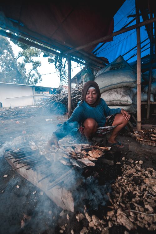 Free Woman grilling fish outdoors in traditional Indonesian setting, surrounded by smoke. Stock Photo