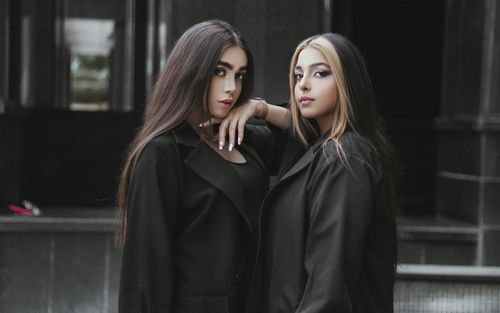 Free Two fashionable women in black coats pose with confidence against a dark, urban backdrop. Modern and chic look. Stock Photo