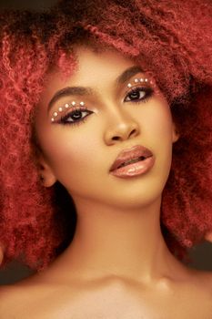 Close-up portrait of a beautiful woman with pink curly hair, artistic makeup, and shimmering details.