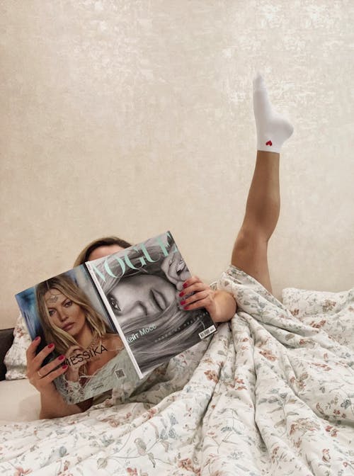 Woman Raising Her Foot While Holding Magazine on Bed 