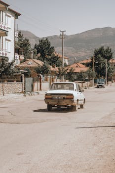 A classic car drives down a quiet urban street surrounded by houses, captured in a vintage style.