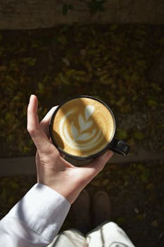 A person holding a coffee cup with intricate latte art, surrounded by leaves outdoors, viewed from above.