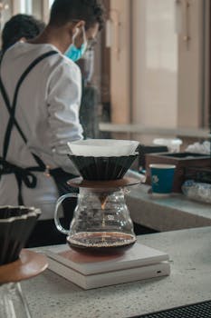 A barista prepares pour-over coffee at a café, highlighting a brewing setup on a counter.