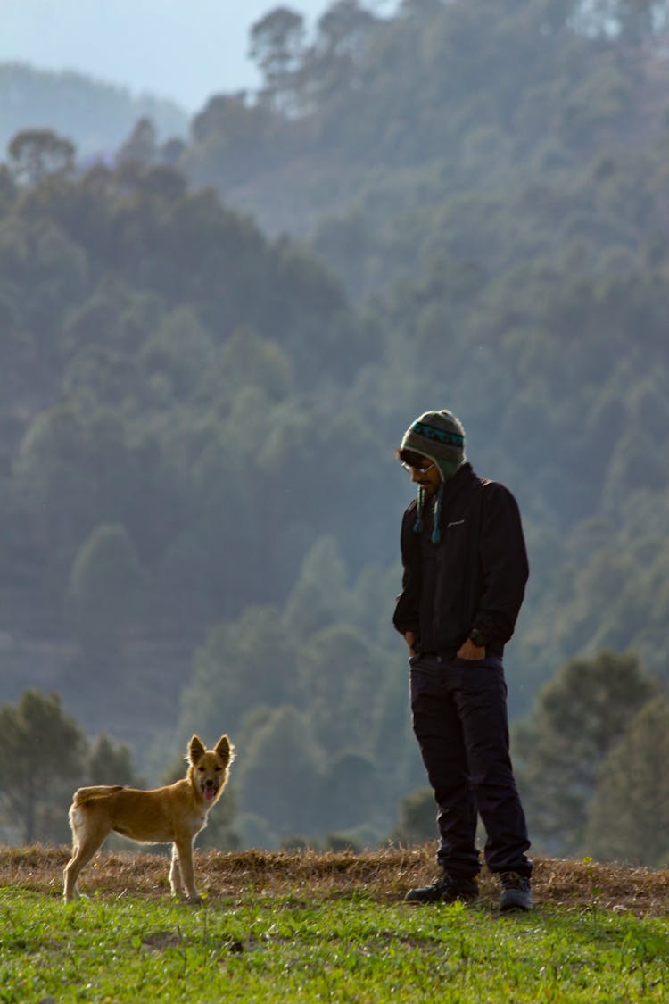 Man In Black Jacket And Pants Standing Beside Brown Dog On Grass Field 