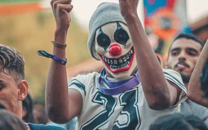 Energetic crowd enjoying an outdoor concert with a masked participant raising hands in excitement.