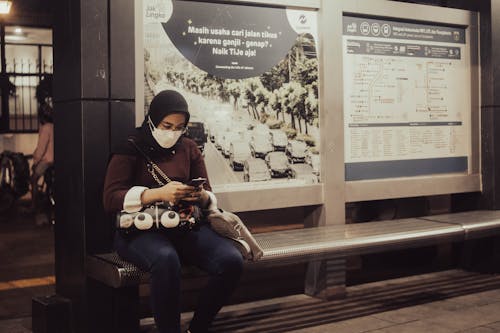 Woman in Black Hijab and Brown Long Sleeve Shirt Sitting on Bench While Using a Phone 