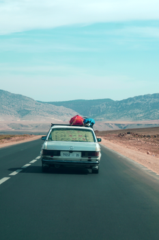 A vintage car travels down a deserted road with mountains in the background under a clear sky.