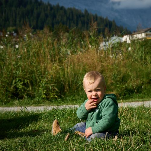 Free A young child in a green hoodie sits on grass with mountains in the background, enjoying a sunny day. Stock Photo