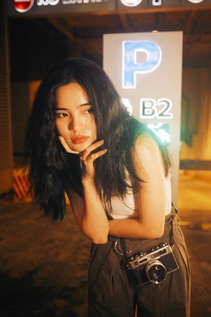 A woman poses stylishly indoors, holding a vintage camera, illuminated by warm lighting.