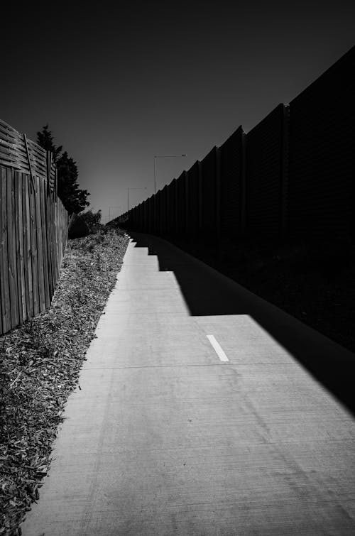 Grayscale Photo of Concrete Road Between Wooden Fence and Concrete Wall