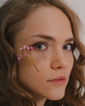 Close-up portrait of a woman with flowers secured by a band-aid under her eye, exploring beauty and vulnerability.