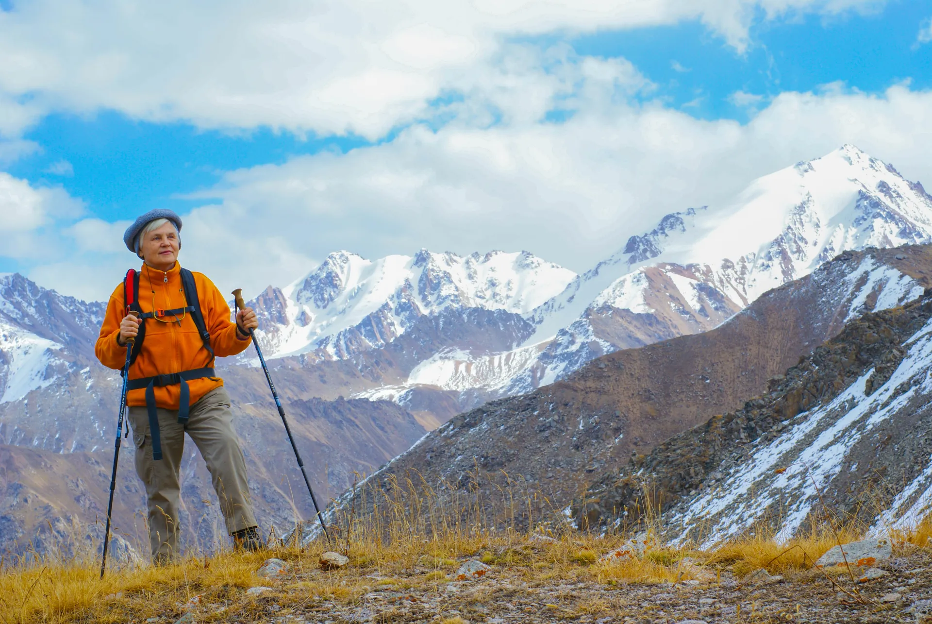 Older woman hiking on a mountain trail with snow-capped peaks behind her Older woman hiking on a mountain trail with snow-capped peaks behind her